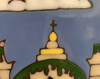 May include: A ceramic tile with a hand-painted design of a building with a green roof and a yellow dome. The building has a blue and white eye-like design on the front. A white cloud with a pink outline is in the upper left corner of the tile.