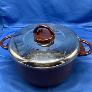 May include: A purple enameled pot with a stainless steel lid and brown plastic handles. The pot has a round shape and a slightly flared rim.