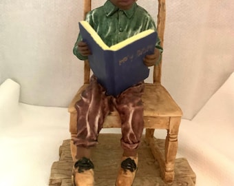 May include: A figurine of a young boy sitting on a wooden chair, reading a blue book with the words "My Bible" on the cover. The boy is wearing a green shirt, brown pants, and brown shoes.