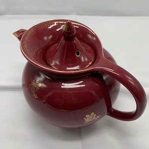May include: A maroon teapot with a gold trim and floral design. The teapot has a lid with a small knob on top.