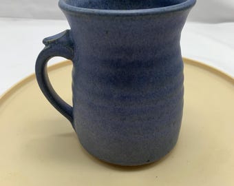 May include: A blue ceramic mug with a speckled finish and a textured, ribbed body. The mug features a curved handle attached to the side and a flared rim. The surface of the mug displays variations in blue tones, creating a mottled appearance.