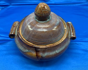 May include: A brown ceramic serving dish with a lid. The dish has a blue and brown glaze with a swirl pattern. The lid has a rounded knob on top.