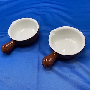 May include: Two small brown ceramic bowls with white interiors and a brown handle. The bowls are on a blue surface.