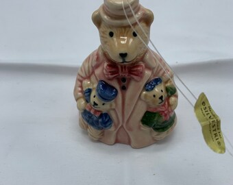 May include: A ceramic bear figurine wearing a pink coat, bow tie, and hat, holding two smaller bear figurines. The bear has a light brown face and is wearing a pink coat with a striped pattern. The figurine has a string for hanging.