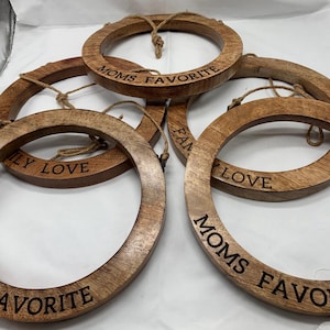 May include: A collection of wooden circular ornaments with engraved text. The ornaments are light brown with a natural wood grain. Some read "MOMS FAVORITE" and "FAMILY LOVE". Each has a jute rope for hanging.