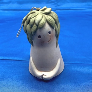 May include: A ceramic bell ornament with a smiling face and green hair. The bell has a white body with a gold string for hanging. The face has black eyes and rosy cheeks. The ornament is on a blue background.