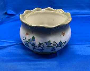 May include: A white ceramic planter with a green rim and a floral design featuring blue flowers and orange butterflies. The planter has a scalloped edge.