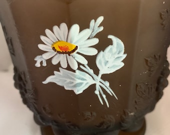 May include: A brown glass vase with a white daisy painted on the side. The vase has a textured, floral design and a scalloped base.
