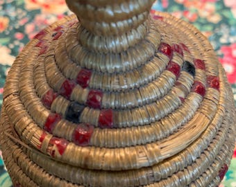 May include: A small, round, woven basket with a lid. The basket is made of natural fibers and has a brown and red pattern.