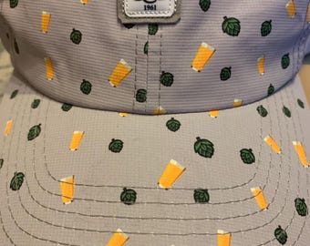 May include: A grey baseball cap with a white patch that says "MCC 1961". The cap has a pattern of green hops and yellow beer glasses.