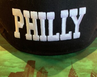 May include: A black baseball cap featuring the word PHILLY embroidered in large white block letters across the front panel. The cap includes small green embroidered eyelets at the top and a structured crown design.