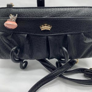 May include: Black crossbody bag with a textured finish. Features a gold-colored crown emblem and a decorative charm. Includes a black shoulder strap and a handle. The bag has a pleated design.
