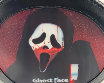 May include: Black backpack with a graphic of the Ghost Face mask from the Scream horror film series. The mask is white with red blood details against a red background. The words "Ghost Face" are printed in white.