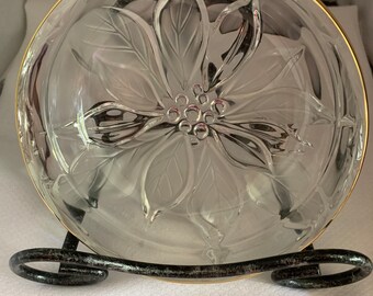 May include: Clear glass bowl with a poinsettia design and a gold rim. The bowl is displayed on a black metal stand. The poinsettia design is etched into the glass, creating a textured effect. The bowl is likely used for serving or display.