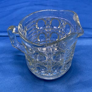 May include: Clear glass creamer with a handle and a spout. The creamer has a geometric pattern etched into the glass. The creamer is sitting on a blue surface.