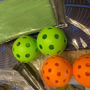 May include: A set of four pickleball balls, two green and two orange, with holes throughout the surface. The balls are packaged with a green mesh towel and a black and blue pickleball paddle.