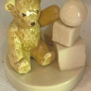 May include: A ceramic figurine of a yellow teddy bear sitting and playing with a stack of three white blocks. The bear has a brown nose and black eyes.