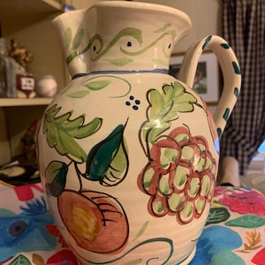 May include: White ceramic pitcher with a hand-painted design of green leaves, red grapes, and a peach. The pitcher has a blue and green handle.