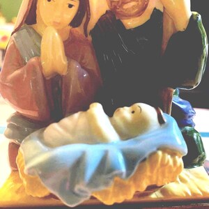 May include: A ceramic nativity scene featuring Mary, Joseph, and baby Jesus. Mary is in a red robe with hands clasped in prayer. Joseph is in a black robe. Baby Jesus is in a blue blanket in a manger. The figures are painted with soft colors.