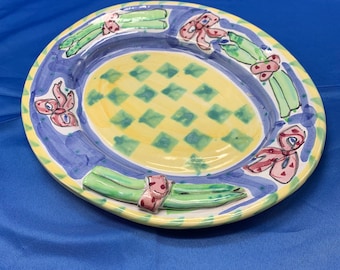 May include: A ceramic serving plate with a yellow and green checkered pattern and a blue rim. The plate is decorated with green asparagus stalks tied with pink bows.
