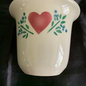 May include: White ceramic vase with a pink heart and green leaves with blue berries.
