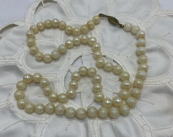 May include: An off-white pearl necklace with a gold clasp, displayed on a white, lace doily. The necklace is composed of many small, round pearls. The clasp is a small, rectangular shape with an ornate pattern.