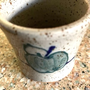 May include: A small, speckled ceramic pot with a wide opening. The pot is off-white with brown speckles and a hand-painted apple design in shades of blue and green. The pot is likely used for holding small items or as a decorative piece.