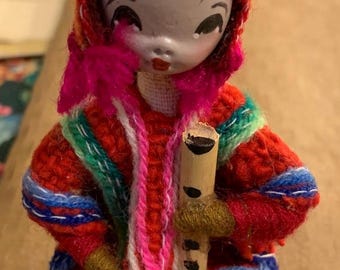 May include: A small, hand-crafted doll wearing a colorful, crocheted outfit and holding a wooden flute. The doll has a painted face with black hair and blue eyes.