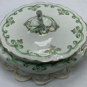 May include: A white porcelain serving dish with a lid, adorned with green and gold floral patterns. The dish has two handles and a decorative knob on the lid. The rim of the dish and lid are scalloped.