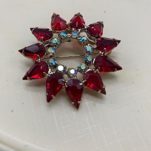 May include: A vintage brooch with a sunburst design. The brooch features teardrop-shaped, faceted red stones set in a gold-tone metal frame. Small, iridescent crystals are interspersed within the design, adding sparkle.