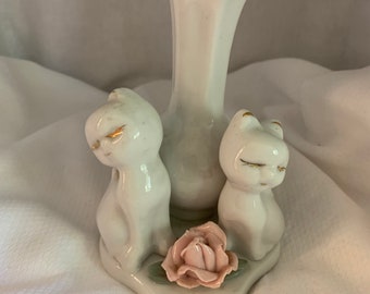 May include: White porcelain vase with two white porcelain cats on either side of the vase. The vase has a scalloped rim and the cats have gold accents. A pink rose is in front of the vase.