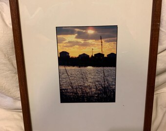 May include: Framed photograph of a sunset over a body of water with a row of houses in the distance. The sky is a vibrant orange and yellow, with clouds reflecting the light. The water is calm and still, with tall grasses in the foreground.