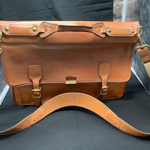 May include: A brown leather messenger bag with a tan canvas shoulder strap. The bag features a top handle, brass hardware, and a front flap with buckle closures. The bag has a rectangular shape and is designed for carrying items.