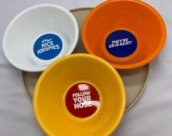 May include: Three round cereal bowls in white, orange, and yellow. The white bowl features a blue logo reading Kellogg's RICE KRISPIES. The orange bowl has a blue centre with the text THEY'RE GR-R-REAT! The yellow bowl has a red centre with FOLLOW YOUR NOSE.