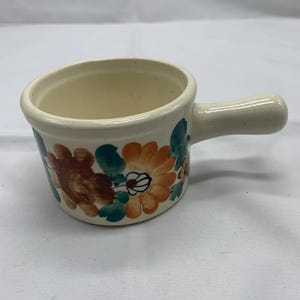 May include: Cream-colored ceramic soup bowl with a handle, adorned with hand-painted floral designs in orange, brown, and teal. The bowl has a slightly angled rim and a sturdy handle for easy use. A charming addition to any kitchen.