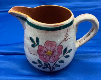 May include: A white ceramic pitcher with a brown rim and a green and brown floral design. The pitcher has a pink flower with a yellow center and green leaves.