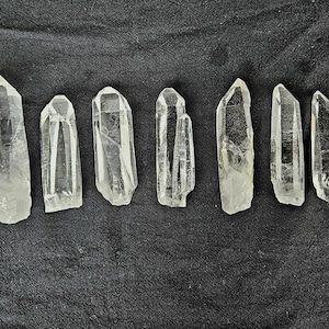 以下が含まれることがあります： さまざまなサイズの7つの透明な水晶ポイントが、暗く質感のある表面に配置されています。結晶は半透明で、内部構造と自然な形成が見えます。結晶は明るくニュートラルな色です。