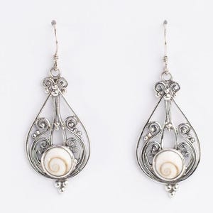 May include: A pair of silver dangle earrings featuring an intricate wirework teardrop frame with decorative scrolls and small silver beads. Each earring centers a round, cream-colored shiva eye shell with a natural spiral pattern at the base.