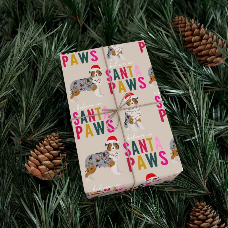 Op de afbeelding: Beige inpakpapier met een herhalend patroon van Australian Shepherd honden die kerstverlichting dragen en de tekst "Geloof in Sinterklaas Pootjes".