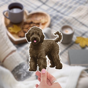 K&ouml;nnte beinhalten: Ein brauner Hundeaufkleber mit lockigem Fell und rosa Zunge. Der Aufkleber wird von einer Hand mit Ringen gehalten. Der Hund steht mit wei&szlig;em Umriss. Der Hintergrund ist verschwommen, mit einer Tasse Tee und Keksen.