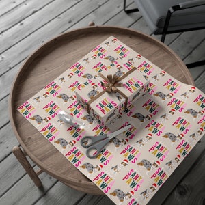 Op de afbeelding: Een cadeau ingepakt in bruin cadeaupapier met een herhalend patroon van blauwe en witte honden met kerstmanmutsen en de tekst "Santa Paws". Het cadeau is vastgebonden met een bruin lint en strik.