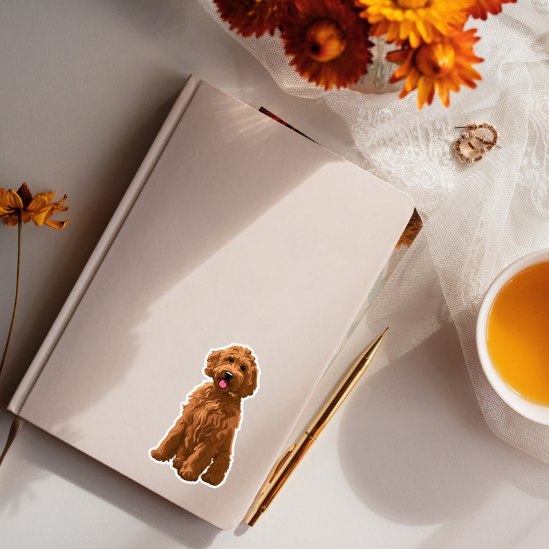 Puede incluir: Una pegatina de perro doodle marr&oacute;n con la lengua rosa que sobresale. La pegatina est&aacute; en una libreta blanca con un bol&iacute;grafo dorado y una taza de t&eacute; en el fondo.