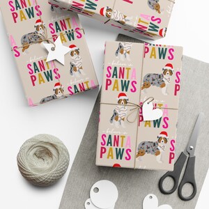 Op de afbeelding: Inpakpapier met een blauwe merle Australian Shepherd-hond die een kerstmanmuts draagt en omringd is door de tekst "I believe in Santa Paws".
