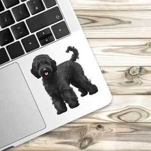 K&ouml;nnte beinhalten: Aufkleber eines schwarzen Portugiesischen Wasserhundes mit lockigem Fell und rosa Zunge, auf einem Laptop. Der Aufkleber befindet sich auf der Ecke des Laptops, mit einer Holzoberfl&auml;che im Hintergrund.