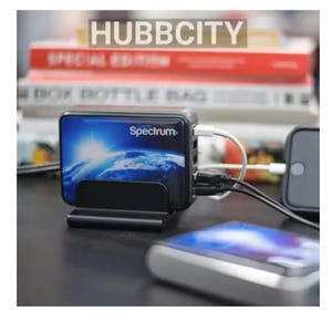 May include: A black Spectrum hub with multiple ports, connected to various devices via white and black cables. The hub is in a black stand, with the word "Spectrum" visible. The background includes books and the text "HUBBCITY".