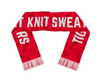 May include: A red knit scarf with white text that reads "T KNIT SWEAT TIG RS". The scarf has white fringe at the ends and a subtle leaf pattern. This winter accessory is perfect for staying warm.