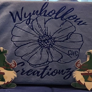 May include: A blue hoodie with the text "Wynhollow Creations" and a large flower design. Two green gnome ornaments with reindeer antlers are in front of the hoodie.