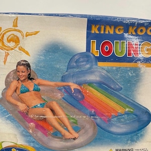 Könnte beinhalten: Karton für einen aufblasbaren Pool-Schwimmkörper von King Koil Lounge. Der Schwimmkörper ist transparent mit Regenbogenfarben und misst 160 cm x 85 cm. Die Box zeigt eine Frau im Bikini, die auf dem Schwimmkörper liegt. Der Text "THE WET SET" ist sichtbar.