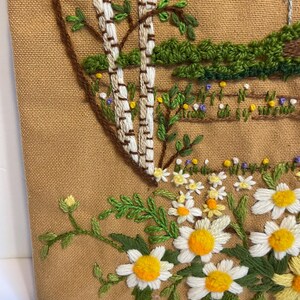 May include: Embroidered wall hanging with a pastoral scene. It shows white birch trees, green foliage, and a field of daisies with yellow centres. The background is a tan fabric.
