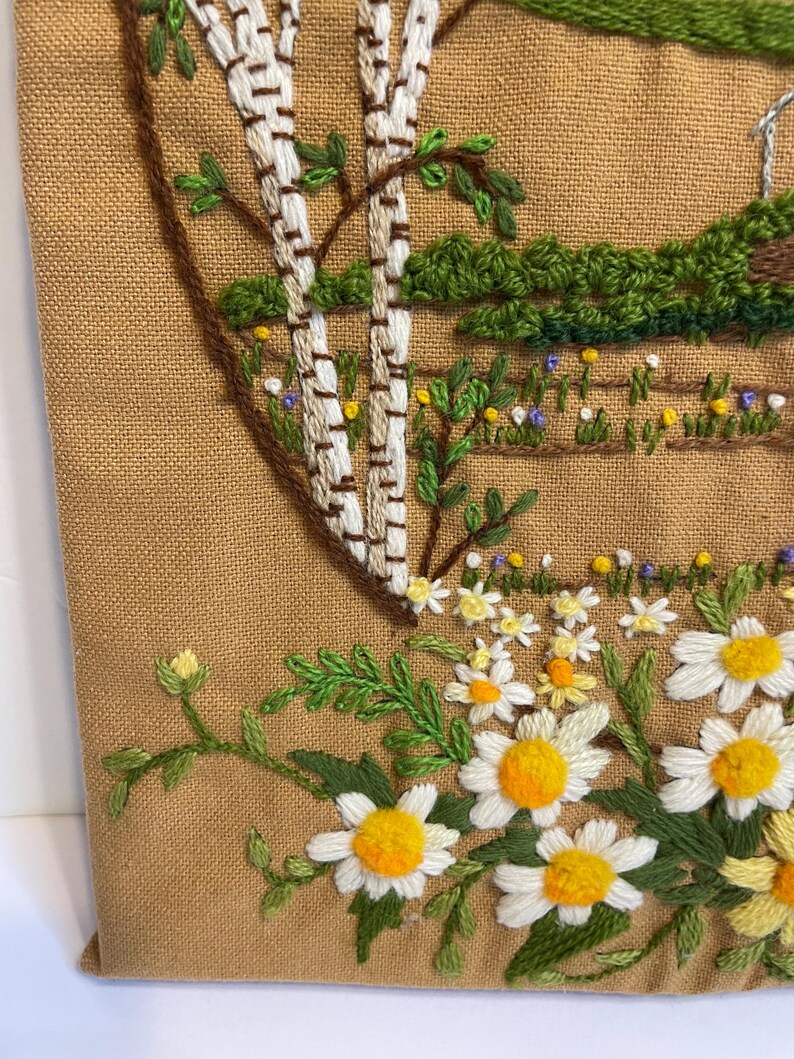 May include: Embroidered wall hanging with a pastoral scene. It shows white birch trees, green foliage, and a field of daisies with yellow centres. The background is a tan fabric.
